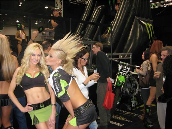 Women in Monster Energy outfits pose in the foreground while attendees stand near a green Kawasaki dirt bike and black inflatable structures with a Monster logo.