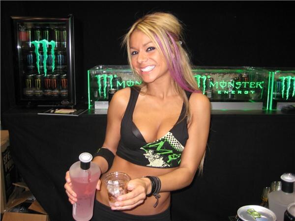 A smiling woman with blonde and purple hair holds a bottle of pink liquid and a glass of ice at a bar with glowing green Monster Energy signs and a cooler of cans behind her.