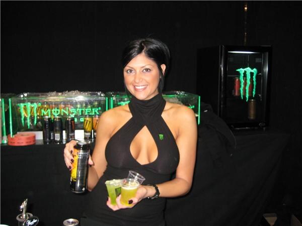 A smiling dark-haired woman in a black halter top holds an energy drink can and two small cups of green drink in front of glowing Monster Energy signs.