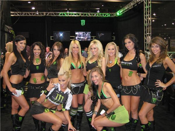 A group of women in black and green Monster Energy outfits pose together in front of a dark booth with a green Monster logo and video screens overhead.