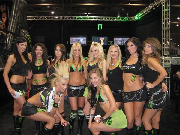 Eight women in black and green Monster Energy outfits pose close together in a booth, one crouching in front, with a dark backdrop and screens behind them.