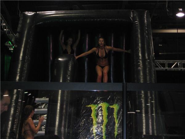Two women in black bikinis stand at the top of a black inflatable slide with a green Monster logo on its front, while others stand nearby below.