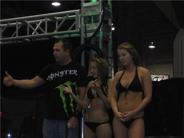 A man in a black Monster T-shirt gestures with a thumb while two women in black bikinis with wet hair stand beside him near a metal truss.