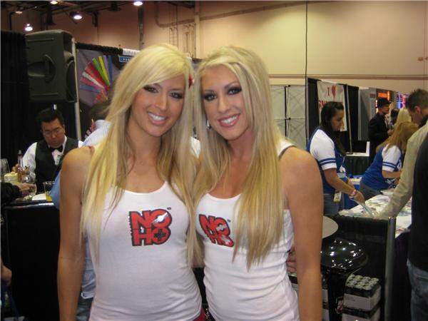 Two smiling blonde women in white NOHO tank tops stand side by side in front of a trade show floor with booths and other attendees behind them.