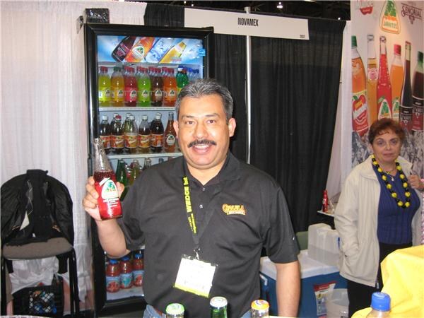 A smiling mustached man holds up a bottle of Jarritos soda beside a cooler of bottled sodas under a Novamex sign, with a woman standing behind him.