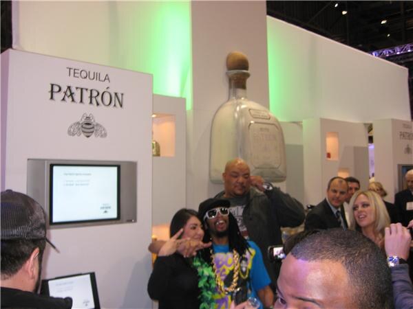 A man in sunglasses and chains poses with a woman at the Patrón tequila display, which has a large Patrón bottle, while other attendees stand around.