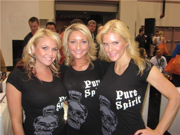 Three smiling women with blonde hair wear black Pure Spirit T-shirts with skull graphics, with other attendees standing behind them.