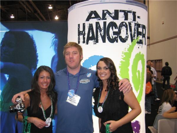 A man in a blue polo shirt stands between two smiling women in black outfits in front of a tall white column reading Anti-Hangover. They wear bead necklaces and badges; one woman holds a camera.