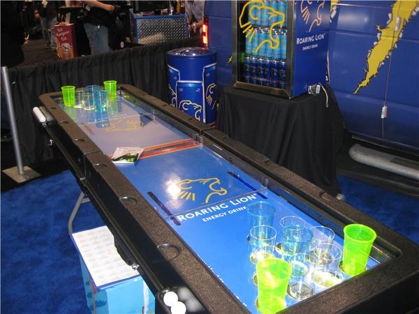 A black and blue beer pong table printed with the Roaring Lion Energy Drink logo, with green and clear cups set at both ends. A lit Roaring Lion can cooler stands on a black-draped table behind it.