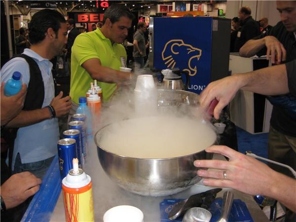 A man in a lime green polo works at a booth counter beside a large steel bowl full of white fog. Roaring Lion cans, spray bottles and a lion logo cooler are nearby, with onlookers around.