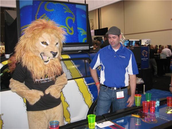 A person in a lion mascot costume wearing a Roaring Lion shirt stands beside a man in a blue polo and gray cap. A beer pong table with colored cups is in front of them.