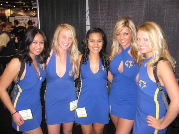 Five smiling women in matching blue dresses with a gold lion emblem and yellow trim pose together, wearing badges and backpacks, in a trade show hall.