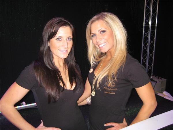 Two smiling women in black shirts pose together against a dark backdrop. The woman on the right, with long blonde hair, wears a Tito's Handmade Vodka shirt.