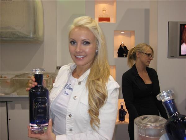 A smiling blonde woman in a white jacket holds up a dark blue bottle of Ultimat Vodka. A woman in black with glasses stands behind her near lit shelves holding bottles.