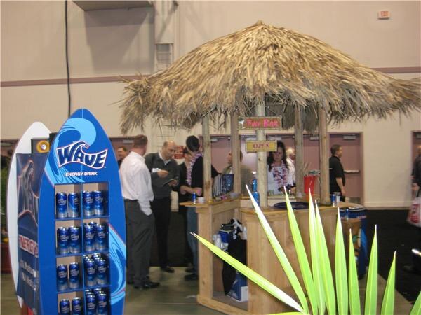 Wave Energy Drink booth with a thatched tiki hut bar and a blue surfboard-shaped display of Wave cans in the foreground. Several people stand and talk near the hut.