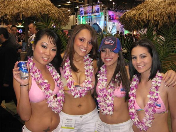 Four smiling women in flower leis and pink tops pose arm in arm under thatched roofs. One holds a can of Wave Energy Drink and another wears a Wave cap.