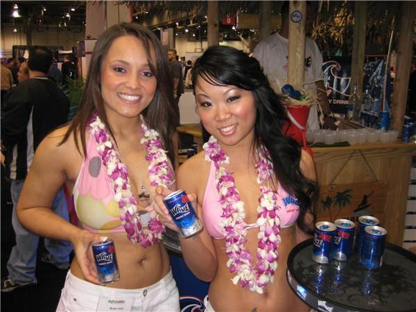 Two smiling women in flower leis and pink tops hold cans of Wave Energy Drink in front of a tiki-style booth with more cans on a round table at right.