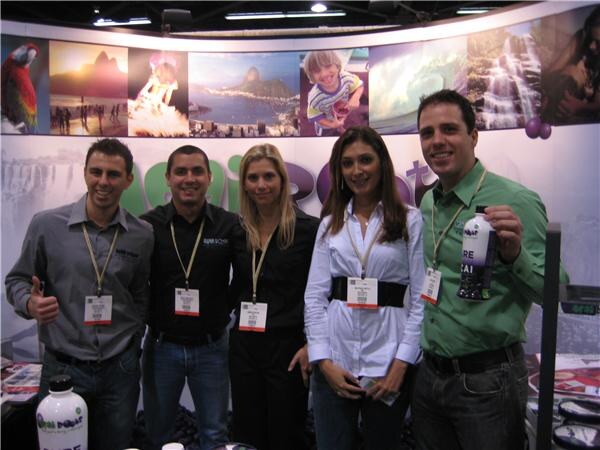 Four men and a woman wearing badges stand together at a booth with scenic photo panels. One man gives a thumbs up and another holds a bottle; Acai Roots bottles sit in front.