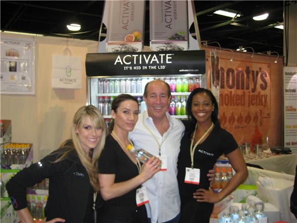 Three women and a man in badges pose in front of an Activate beverage cooler. A Monty's Smoked Jerky banner hangs at right, and bottles are stacked nearby.
