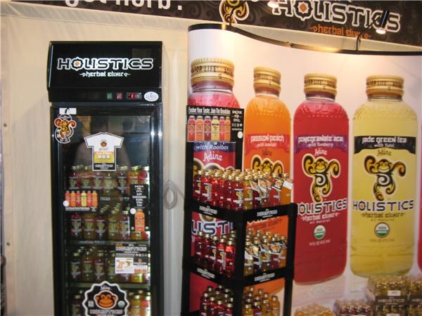 Adina Holistics herbal elixir display with a lit glass-door cooler of bottles, a tiered rack of red bottles and a large banner of passion peach, pomegranate acai and jade green tea bottles.