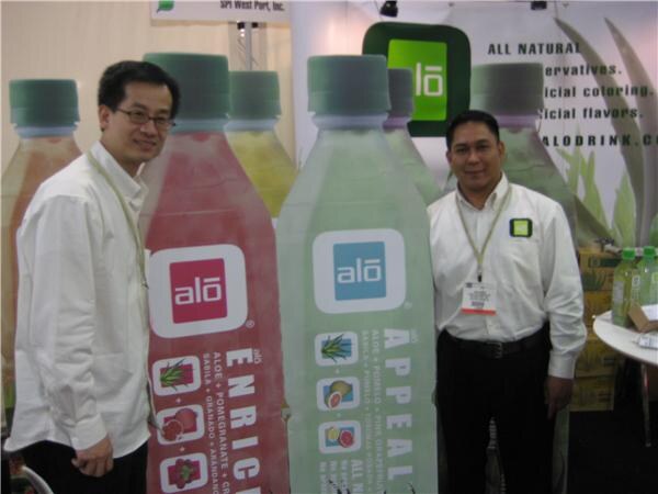 Two men in white shirts stand beside large cutout bottles of Alo drink in red and green. A sign behind them has the Alo logo, and small bottles sit on a table at right.