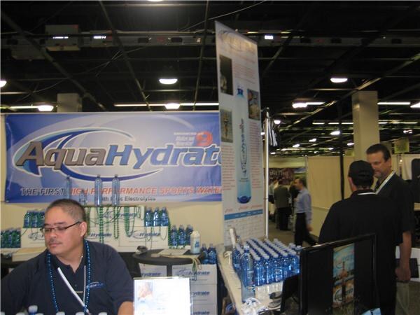 A man in glasses and a dark shirt sits at an AquaHydrate booth with a banner and bottles of water on tables. Two other people stand at right in the aisle.