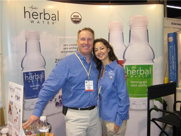 A smiling man and woman in blue shirts stand together in front of Ayala's Herbal Water banners showing lavender mint and lemongrass mint vanilla bottles and a USDA Organic seal.