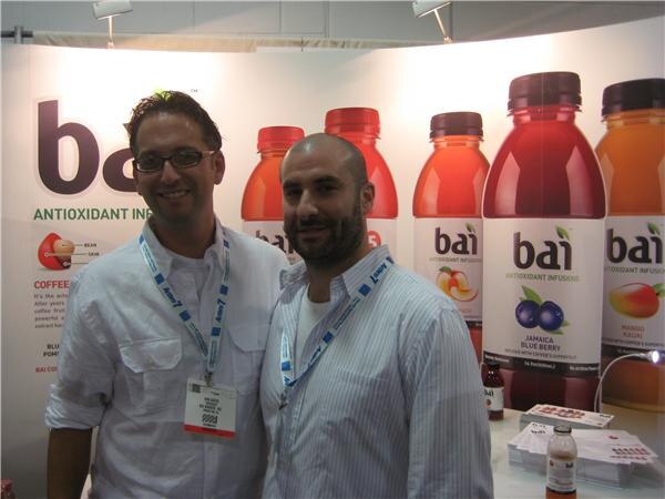Two men with convention lanyards stand side by side in front of a Bai display of antioxidant infusion bottles, including Jamaica Blue Berry and Mango Kauai flavors.