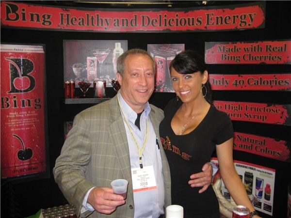 A man in a plaid blazer holding a small sample cup stands with a woman in a black Bing shirt in front of red signs reading Made with Real Bing Cherries and Only 40 Calories.