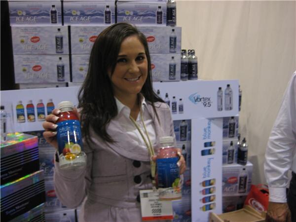 A smiling woman holds two Blue Monkey bottles in front of stacked cases of Ice Age water and a Vortex 9.5 display. Part of another person's white sleeve is visible at right.