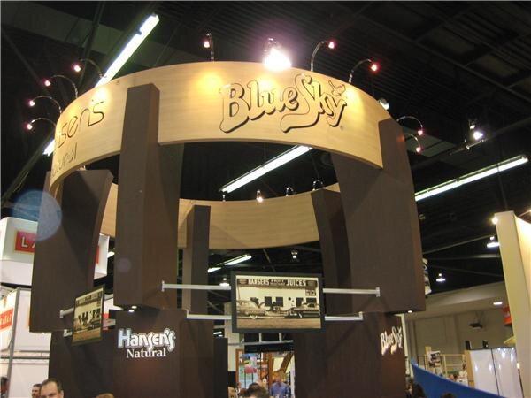A circular wooden booth structure displays Blue Sky signage on top, with Hansen's Natural logos and a vintage photo of a Hansen's juice shop on the lower panels.
