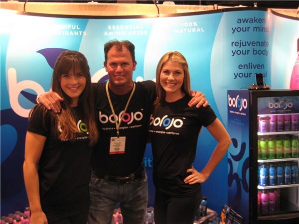 A man in a black Borojo shirt stands with his arms around two smiling women in matching Borojo shirts, with a backdrop reading awaken your mind and a refrigerated case of bottles at right.