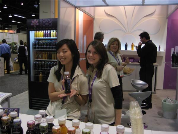 Two smiling young women in cream shirts and purple lanyards stand at a counter of Bossa Nova juice bottles, one holding a bottle, with a lit Bossa Nova cooler behind them.
