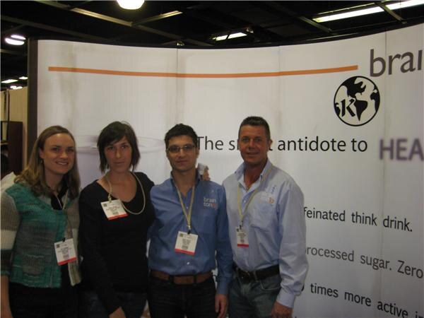 Two women and two men wearing show badges stand in a row in front of a white Brain Toniq banner with partial text about a caffeinated think drink.