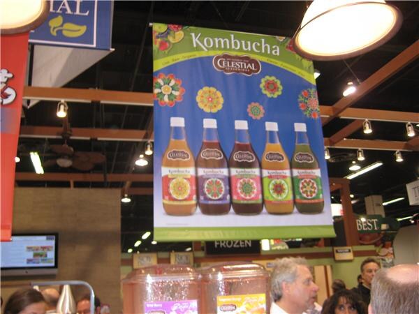 A hanging banner reads Kombucha with the Celestial Seasonings logo and five bottles with colorful labels. Several people stand below it and sample dispensers sit at the bottom.