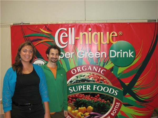 A woman in a blue cardigan and a bearded man in a green jacket stand in front of a red Cell-nique Super Green Drink banner featuring organic super foods.