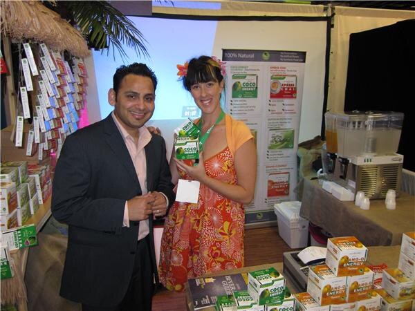A man in a dark suit stands beside a smiling woman with a flower in her hair who holds a Coco Energy box, with more Coco Energy boxes on the table and a drink dispenser behind them.
