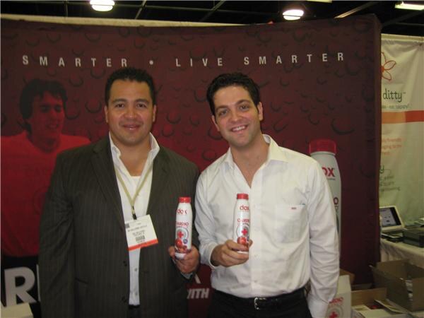 Two men stand together, each holding a bottle of Dox Cardio Water, in front of a red Dox banner reading Smarter, Live Smarter.