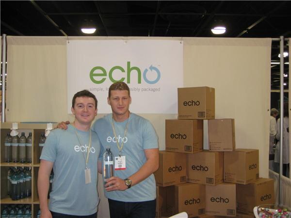 Two men in light blue Echo T-shirts and lanyards stand in the Echo booth, one holding a water bottle, beside stacked Echo cardboard boxes and a shelf of bottles.