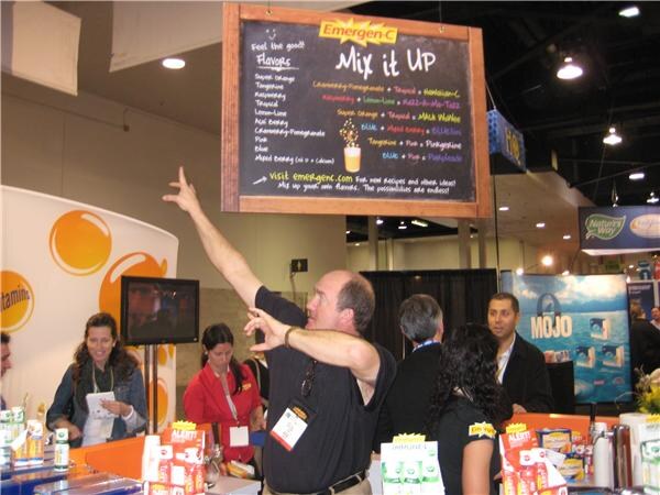 A man in a black shirt points upward toward an Emergen-C "Mix it Up" flavor chalkboard sign while booth staff stand behind. A Mojo banner and a Nature's Way sign are in the background.