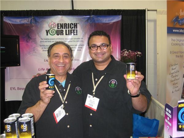 Two smiling men in black shirts and badges each hold up a small DeCaf can in front of an Enrich Your Life! banner, with more cans in the lower corner.