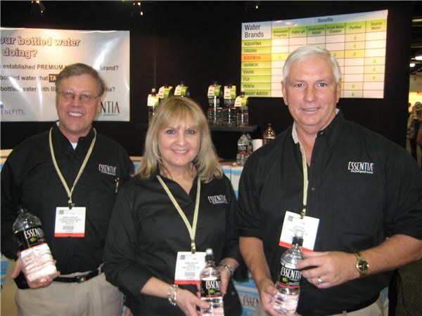Two men and a woman in black Essentia shirts and badges each hold a bottle of Essentia water, with a water brand comparison chart on the wall behind them.