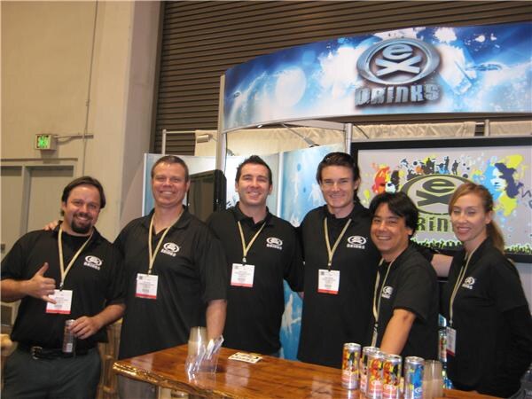 Six people in black eX Drinks shirts and lanyards pose together behind a wooden counter holding cans, in front of an eX Drinks booth sign.