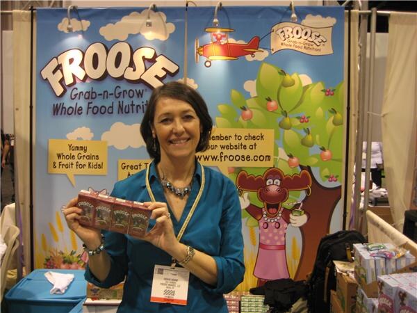 A smiling woman in a teal top and lanyard holds up a few small Froose boxes in front of a colorful Froose backdrop with a cartoon cow and fruit trees.