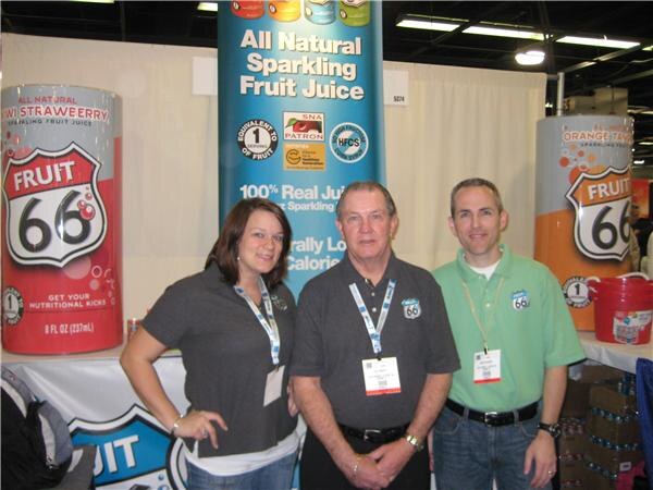 Two men and a woman stand in the Fruit 66 booth in front of tall cans labeled Fruit 66 and a banner reading All Natural Sparkling Fruit Juice.