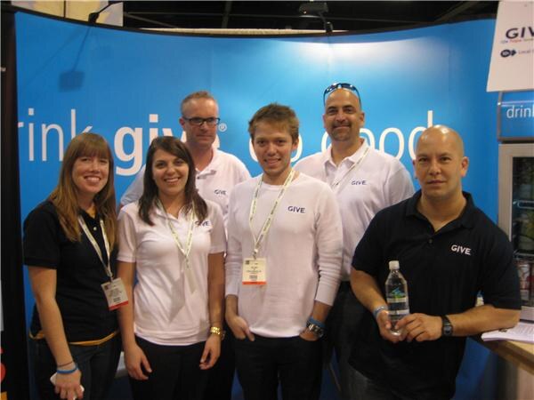 Six people in Give shirts stand together in front of a blue Give banner reading drink give, do good; the man at right holds a bottle of water.