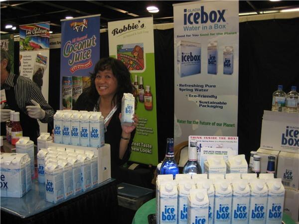 A smiling woman with dark hair holds a Glacia Icebox water carton in a booth. Stacks of Icebox cartons fill the tables, with Icebox and Robbie's banners behind her.