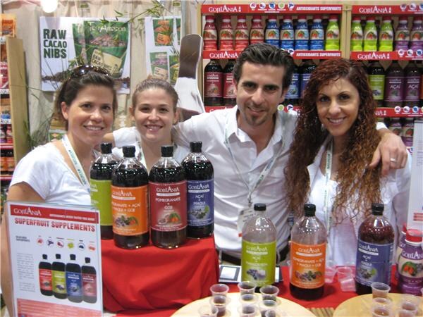 Three women and a man pose with their arms around each other behind a red-draped table of GojiAna superfruit juice bottles, with GojiAna-stocked shelves behind them.