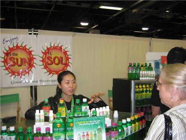 A woman behind a counter of green aloe vera drink bottles gestures as she speaks to an older woman with short light hair. Sun-themed Good Karma Beverage banners hang behind.