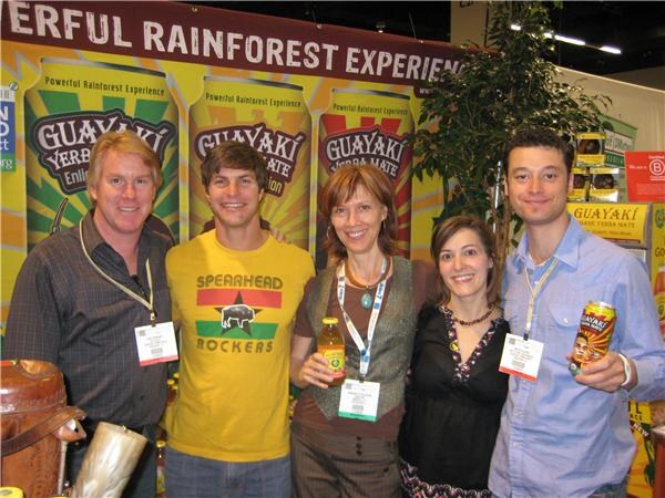 Five people with badges pose together in front of a Guayaki Yerba Mate banner. One man wears a yellow Spearhead Rockers shirt and another holds a Guayaki can.